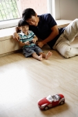 Father and young son sitting floor at home, playing with remote control car - Alex Mares-Manton