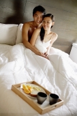 Couple sitting up in bed, looking at camera, breakfast tray at foot of bed - Alex Mares-Manton