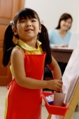 Young girl standing next to easel, smiling at camera, mother in the background - Alex Mares-Manton