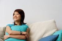 Woman sitting on sofa, hugging cushion, looking away, smiling - Nugene Chiang