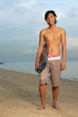 Young man holding skimboard, standing on beach - Yukmin