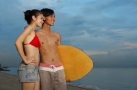 Couple standing on beach, man carrying skimboard, looking away - Yukmin