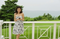 Young woman in floral dress, standing on balcony, holding mobile phone - Yukmin