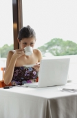 Woman sitting in restaurant, using laptop, drinking from cup - Yukmin