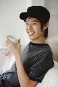 Young man sitting on sofa, holding cup - Yukmin