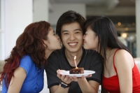 Teenage boy in the middle holding plate with a cake and candle, girls on either side, kissing him on cheek - Yukmin