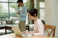Couple at home, woman using laptop, man in the background, on the phone - blueduck