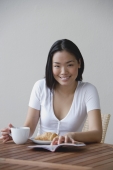 Young woman having breakfast and reading a book, smiling at camera - Yukmin