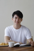 Young man having breakfast and reading a magazine, smiling at camera - Yukmin