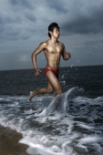 Muscled man wearing swim trunks, running along ocean - Yukmin