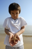 Young boy holding shell on both hands at the beach, looking at camera - Yukmin