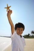 Young boy holding up shell at the beach, looking at camera - Yukmin