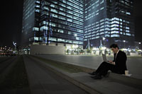 Man sitting on ground working on lap top in front of lit buildings - Alex Mares-Manton