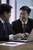 Businessmen smiling at each other while working on laptop - Alex Mares-Manton