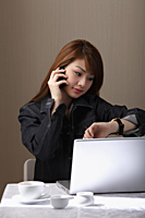 Young woman sitting at table with lap top, talking on phone, looking at watch - Asia Images Group