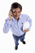 Mature man holding construction hat, using mobile phone, looking at camera - Asia Images Group