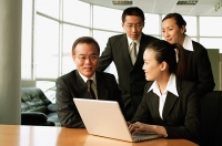 Executives in office, sitting and standing around open laptop, talking - Asia Images Group