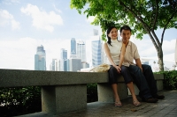 Young couple sitting on park bench, looking at camera, buildings in the background - Asia Images Group