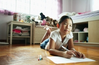 Girl in bedroom, lying on floor, holding pen, looking at camera - Asia Images Group