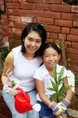 Mother and daughter, sitting in garden, holding plant and watering can - Asia Images Group