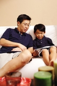 Father and son sitting side by side on sofa, looking at magazine - Asia Images Group