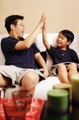 Father and son sitting side by side on sofa, making high five - Asia Images Group