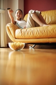 Girl lying on sofa, holding remote control, bowl of popcorn on the floor - Asia Images Group