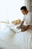 Man sitting on bed, using laptop - Asia Images Group