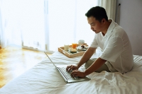 Man sitting on bed, using laptop, breakfast tray next to him - Asia Images Group