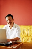 Man sitting on sofa, using laptop, smiling at camera - Asia Images Group