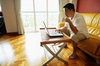 Man sitting in living room, using laptop, drinking from mug - Asia Images Group