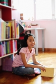 Girl sitting cross-legged on floor, holding book, looking at camera - Asia Images Group