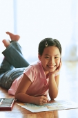 Girl lying on floor, with paper and crayons, looking at camera, hand on chin - Asia Images Group