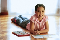 Girl lying on floor, with paper and crayons, looking at camera, smiling - Asia Images Group