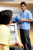 Man standing in bowling ally, holding bowling ball, another man sitting in front of him - Asia Images Group