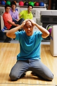 Man kneeling in bowling alley, hands on head, frowning, people in the background watching - Asia Images Group