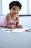 Young girl lying on floor, holding crayons, smiling at camera - Asia Images Group