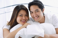 Couple on bed, hugging pillows, smiling at camera, portrait - Asia Images Group