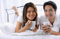 Couple on bed, holding mugs, smiling at camera, portrait - Asia Images Group