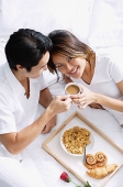 Couple on bed, breakfast tray between them, woman looking at camera - Asia Images Group