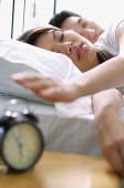 Couple lying on bed, woman reaching to switch of alarm clock in foreground - Asia Images Group