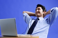 Man wearing headset, using laptop, leaning back, hands behind head - Asia Images Group
