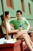 Couple sitting on bench on tennis court, smiling at each other, woman with legs crossed - Asia Images Group