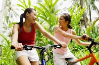 Two girls riding bikes through a park, looking at each other - Asia Images Group