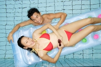 Woman lying on pool raft, man in water next to her, looking up at camera - Asia Images Group