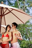 Couple standing under umbrella, holding drinks, looking at each other - Asia Images Group