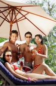 Couples standing under umbrella, holding drinks, one woman sitting on deck chair - Asia Images Group