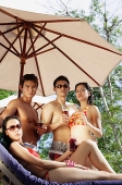 Young adults standing under umbrella, holding drinks, one woman sitting on deck chair - Asia Images Group