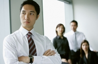 Businessman standing with arms crossed, looking away, people in the background - Asia Images Group