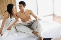 Couple sitting on bed with newspaper, looking at each other - Asia Images Group